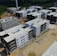 Aerial view of a construction site featuring multiple new, multi-story apartment buildings. Each building has a similar design with a combination of white and dark grey exteriors. The area is surrounded by greenery and suburban residential buildings in the background.